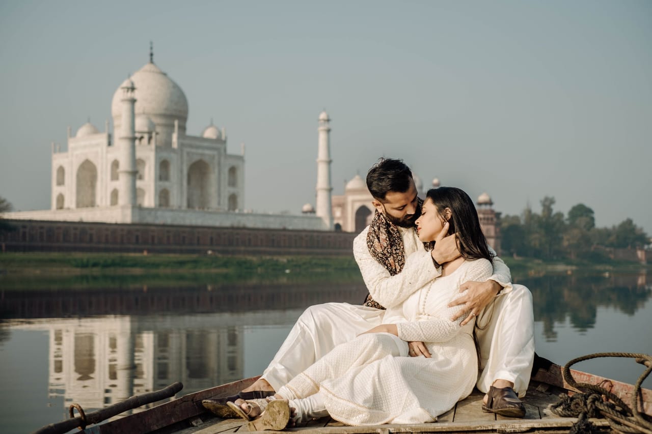 A couple in ivory sit close together on the bow of a wooden boat on the Yamuna, the Taj Mahal and its reflection behind them.