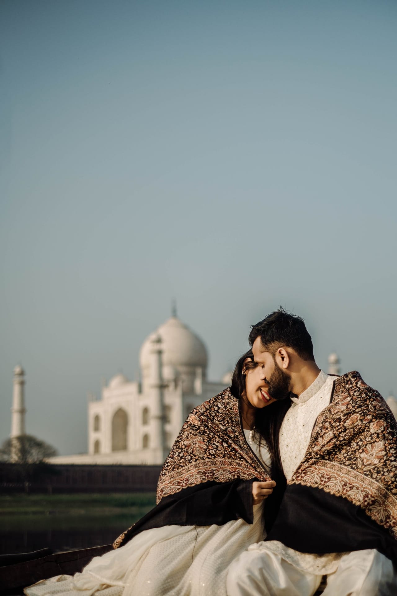 Wrapped together in a dark embroidered shawl, a couple lean into each other with the Taj Mahal softly out of focus behind.