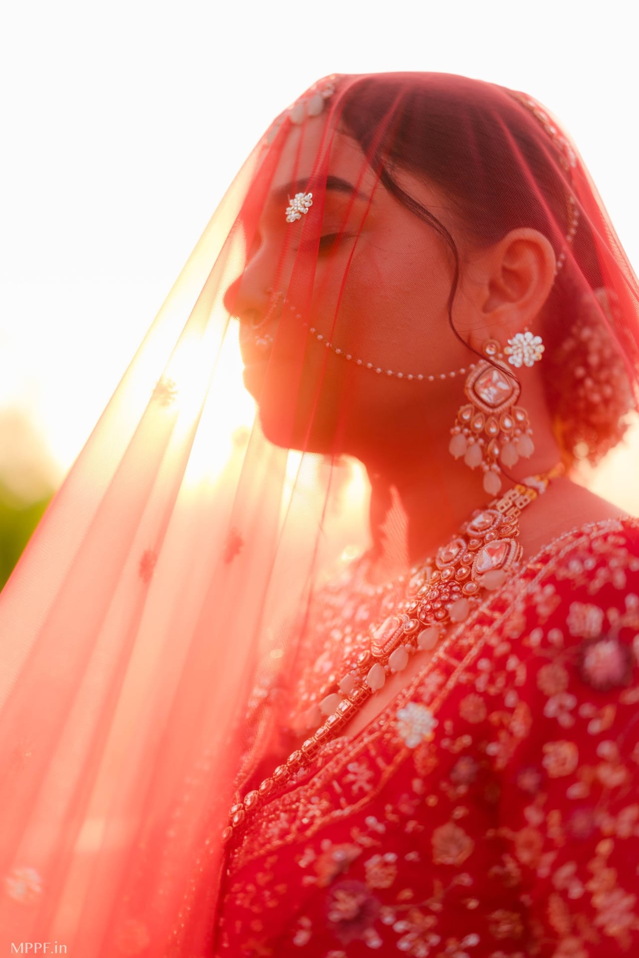 Profile of a bride beneath a sheer red veil, backlit by the sun, wearing large pearl and stone earrings.