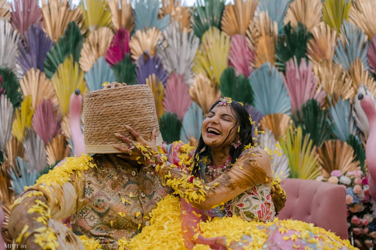 At a haldi in front of a wall of painted palm fans, a bride covered in turmeric laughs as she pushes a woven basket over the groom’s head.