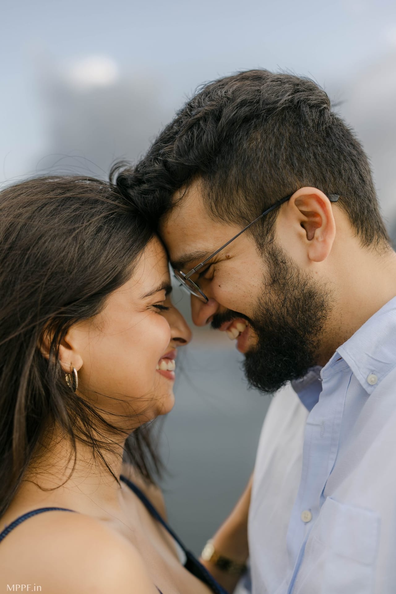 Close portrait of a couple laughing with their foreheads pressed together, grey sky behind.
