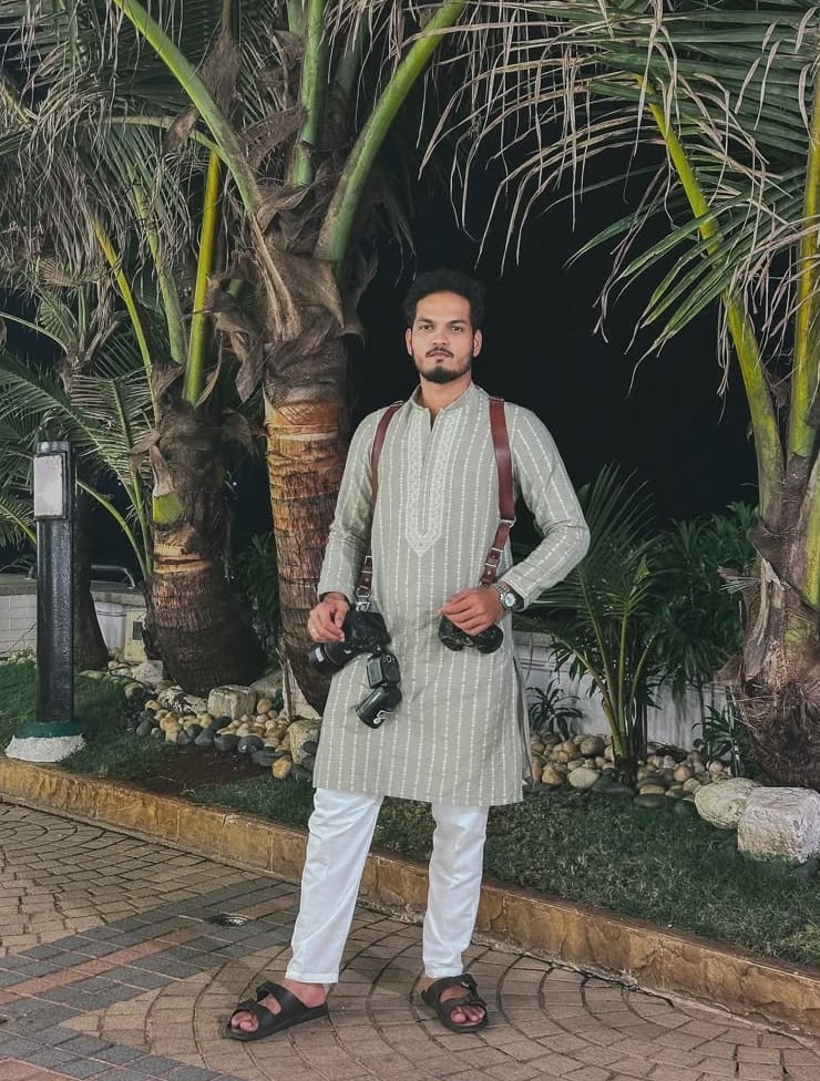 Makrand Parab standing under palm trees at night in a sage kurta, two cameras on a leather harness.