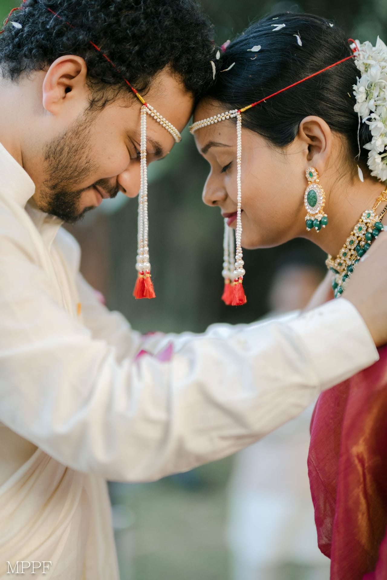 A Maharashtrian couple wearing pearl mundavalya touch foreheads, eyes closed, petals in their hair.