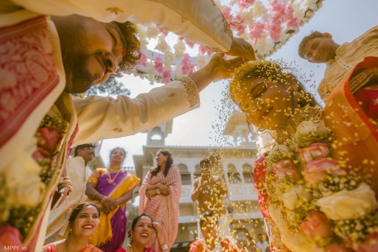 Low-angle view of a groom showering rice over the bride’s head as family members laugh around them in a palace courtyard.