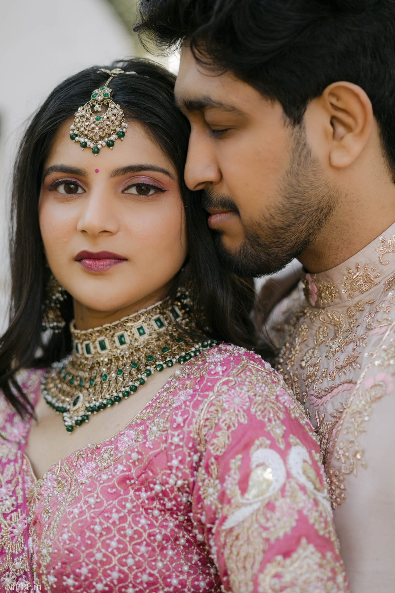 A bride in a pink embroidered blouse and emerald jewellery looks at the camera while the groom rests his face against her head.