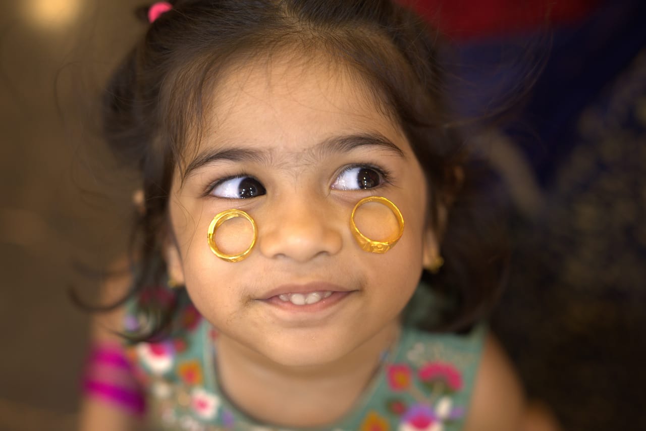 A little girl holds two gold rings against her cheeks and looks sideways with a grin.